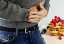 Homem com as mãos sobre o estômago ao lado de um prato de doces, sintoma de gastrite erosiva